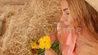 一个年轻漂亮的女孩戴着草帽坐在干草上，手里拿着一朵黄色的花，然后闻了闻。 4k. 4k视频